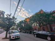 <p>A picture from the Pixel 6a's ultrawide camera, featuring a red building behind some trees in front of a road lined with cars on both sides. In the background is a blue sky and some clouds.</p> 