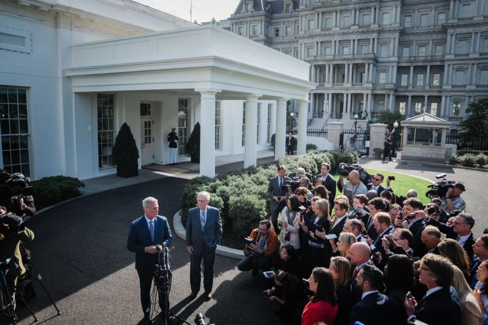 Two men in suits stand near the White House in front of several other people.