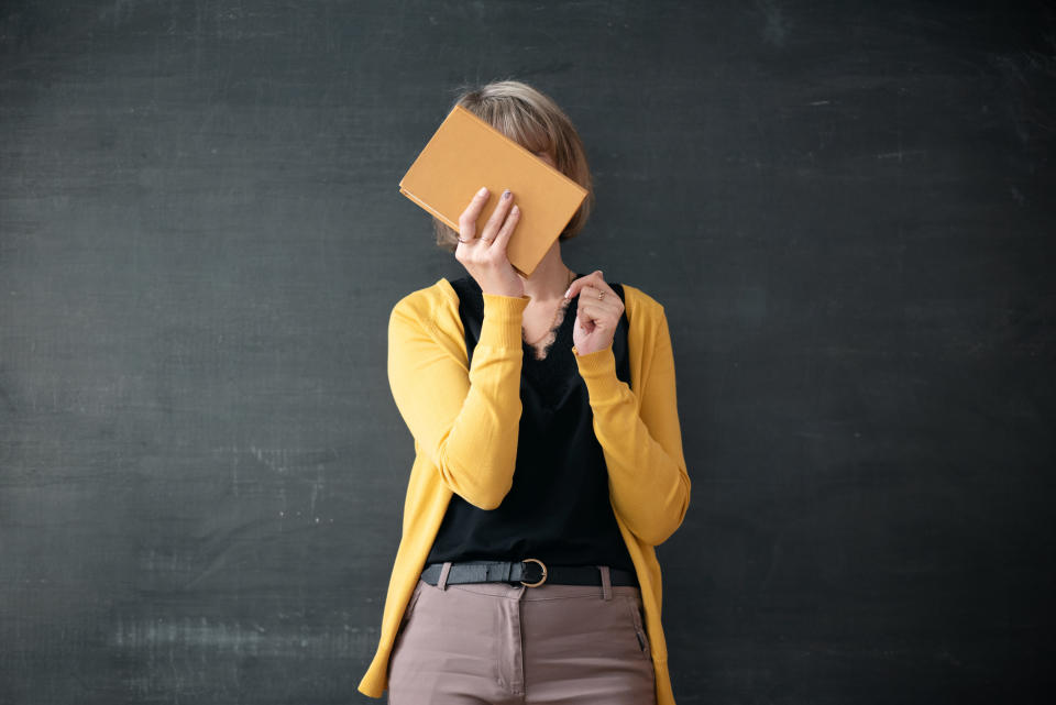 A teacher covering her face with a book in front of the chalkboard