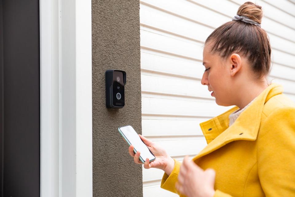 A woman holding a smartphone walks up to a video doorbell.