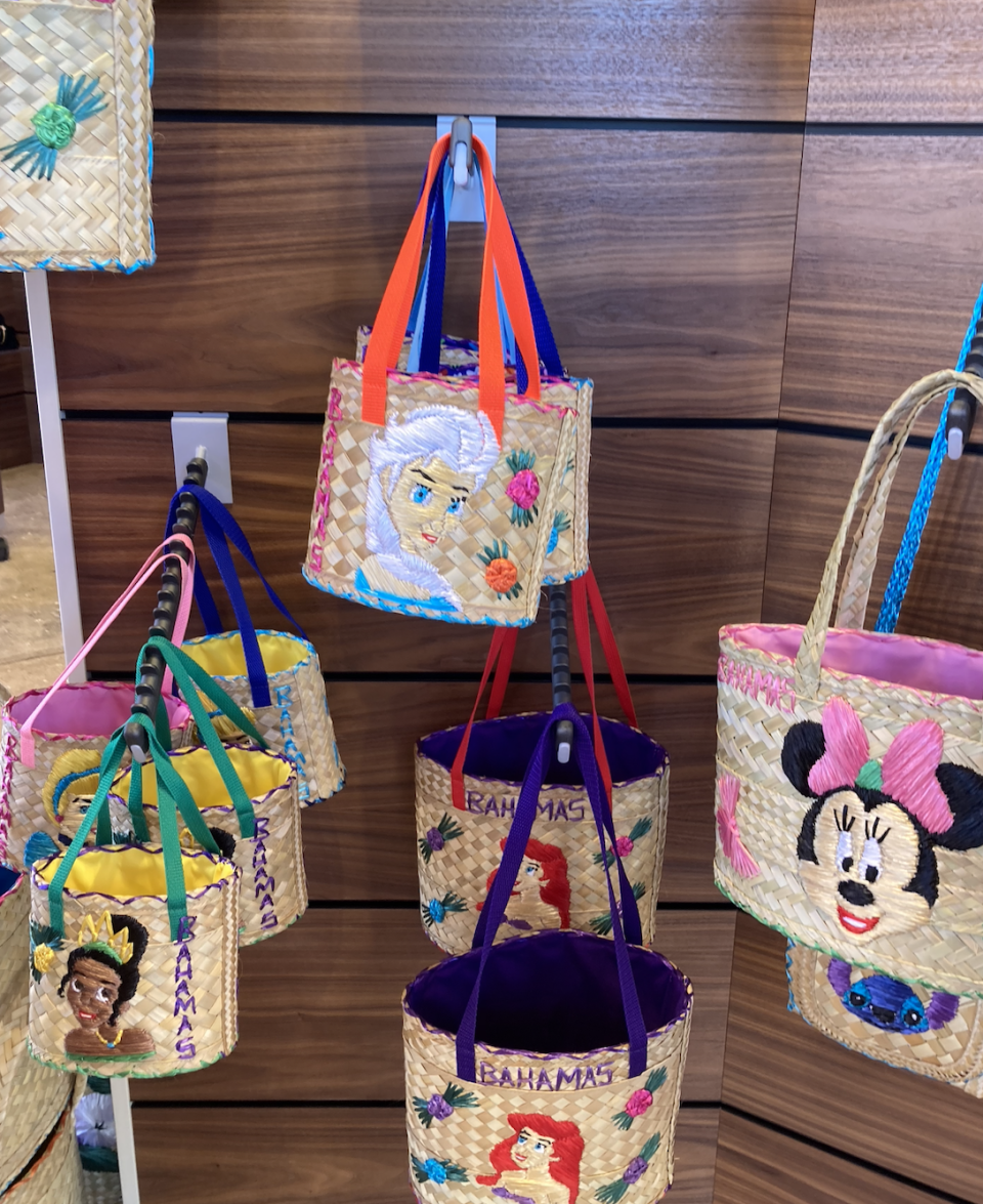 Straw handbags with Disney characters Tiana, Elsa, Ariel, and Minnie Mouse are displayed on wall hooks. Handles are colorful. Bags say "Bahamas."