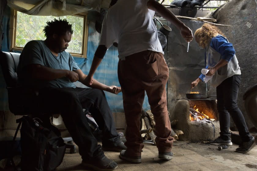 Edward Soto, 27, known as Smokey, shoots up Neiko Clayton, 29, known as Trap, with crystal methamphetamine in Encino, California, on April 29th, 2019. Karla Saenz, 46, who is a mother figure in the homeless community, cooks on an adobe stove. They are in a structure in a homeless community on "The Field," which is an area along the LA River on federal land. The majority of the people who live there use crystal meth.