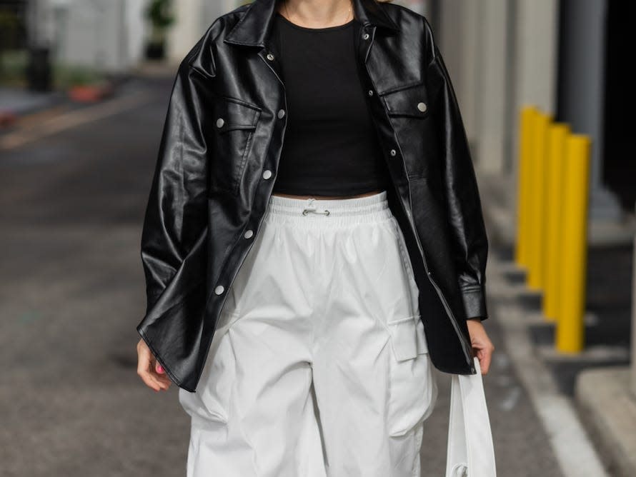 woman walking down a street wearing a black leather jacket over a black t shirt and white wide leg cargo pants