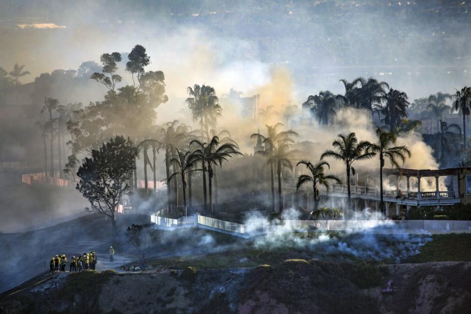 A group of firefighters gather amid clouds of smoke, in front of a burned-out home