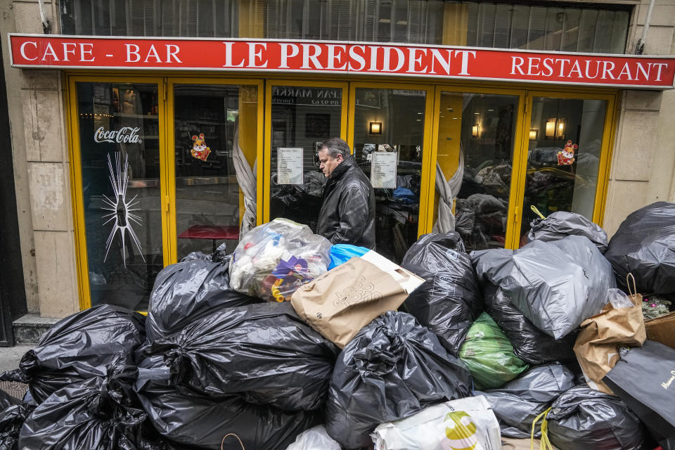 A man walks past an uncollected garbage pile next to the cafe "The President" in Paris, Tuesday, March 21, 2023. The bill pushed through by President Emmanuel Macron without lawmakers' approval still faces a review by the Constitutional Council before it can be signed into law. Meanwhile, oil shipments in the country were disrupted amid strikes at several refineries in western and southern France. (AP Photo/Michel Euler)