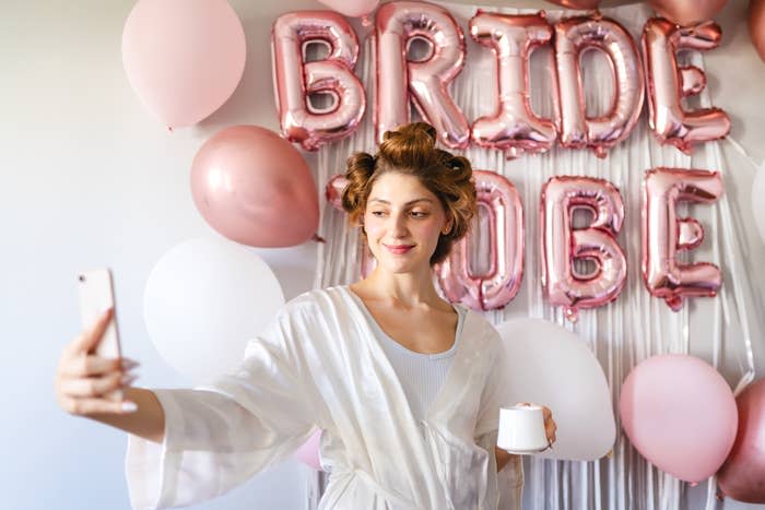 Woman in white robe taking a selfie, with "BRIDE TO BE" balloons in the background