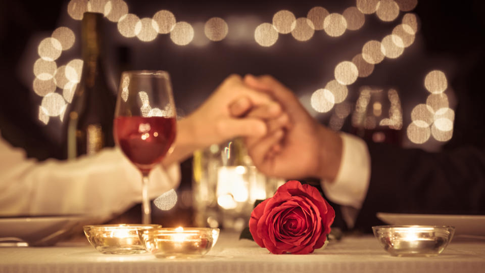 A wine glass and a rose can be seen in the foreground and a couple's clasped hands in the background.