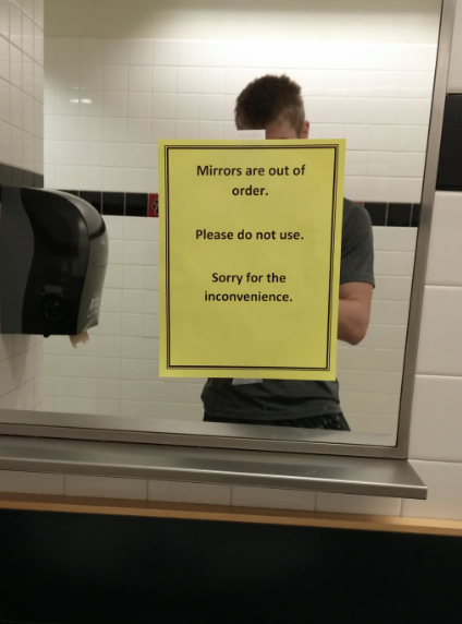 A man stands in a restroom mirror with a sign that reads, "Mirrors are out of order. Please do not use. Sorry for the inconvenience."