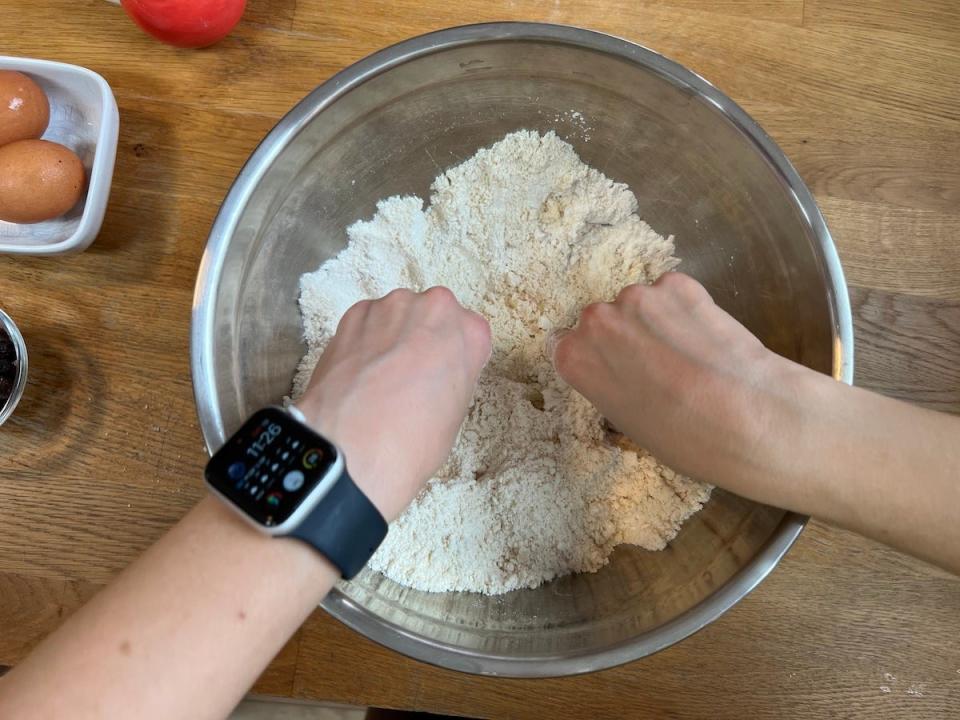 An overhead view of a woman using her fingers to form breadcrumbs out of flour, sugar, butter, and baking powder in a silver mixing bowl.