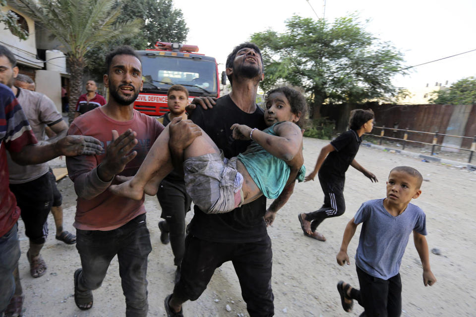 A man, surrounded by other people, carries a girl who is covered in dirt from building rubble along a road (Abed Khaled / AP)