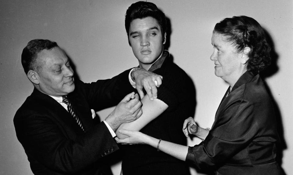 Elvis Presley standing between two medical staff receiving an injection in his arm