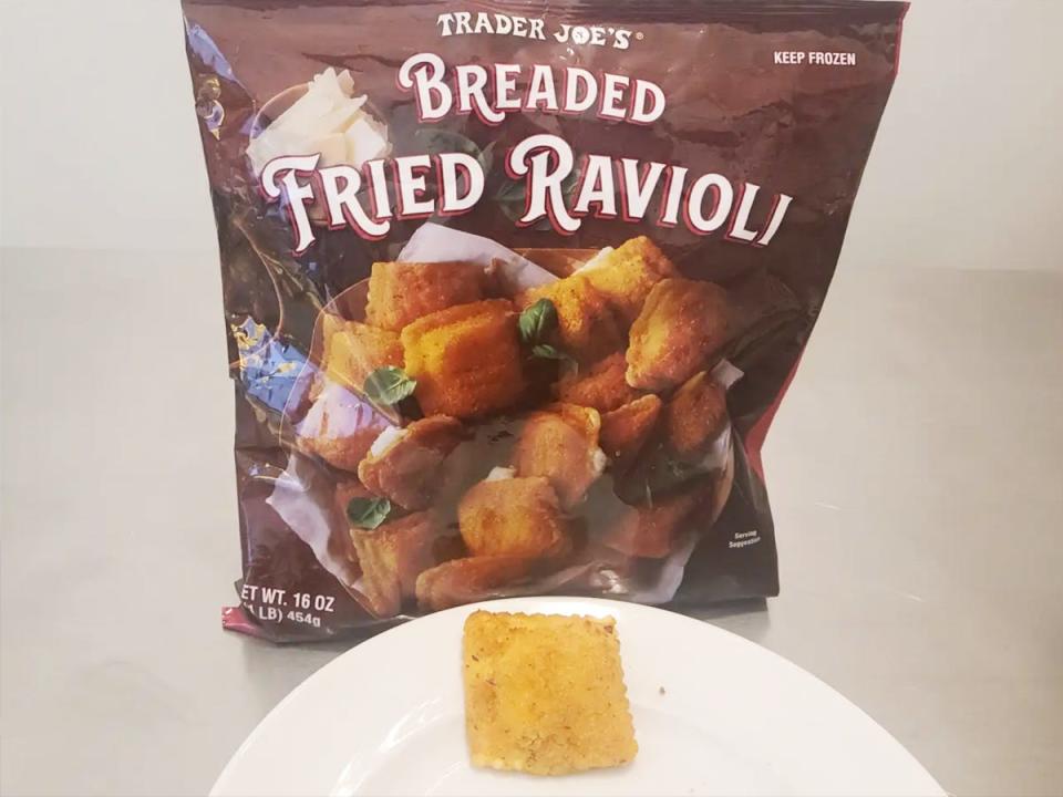 A piece of breaded ravioli sits on a plate with a brown bag of breaded fried ravioli behind plate