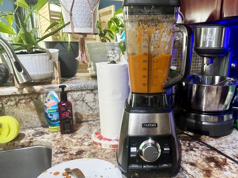 A kitchen counter with a mixer, paper towels, and a blender filled with an orange sauce. Several potted plants sit in the background