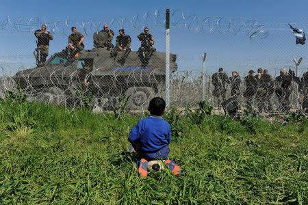 A boy sits on his ball next to a border fence on the Greek side of the border, as Macedonian police stand guard on the Macedonian side, close to a makeshift camp for refugees and migrants near the Greek village of Idomeni, April 12, 2016. Picture taken from the Greek side of the border. REUTERS/Alexandros Avramidis