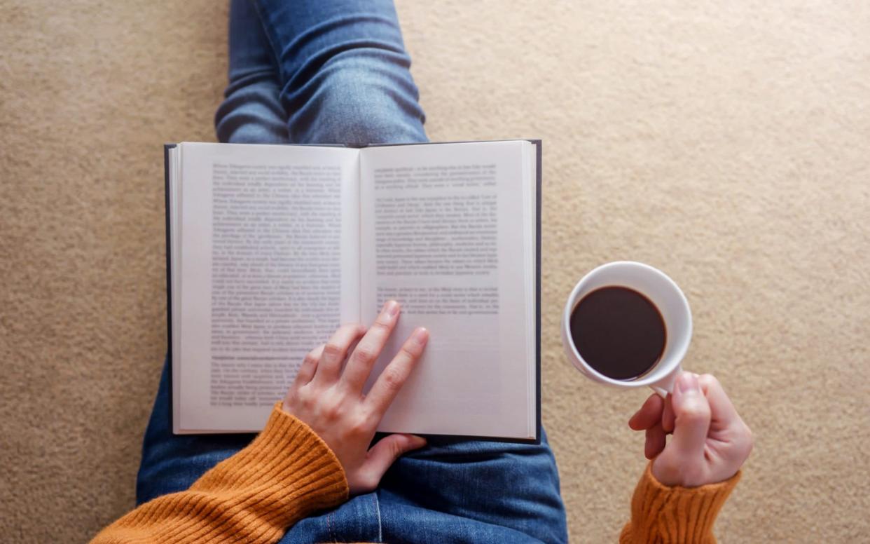 A woman reads a book and holds a mug of coffee - Getty Images Contributor/Sirinapa Wannapat / EyeEm