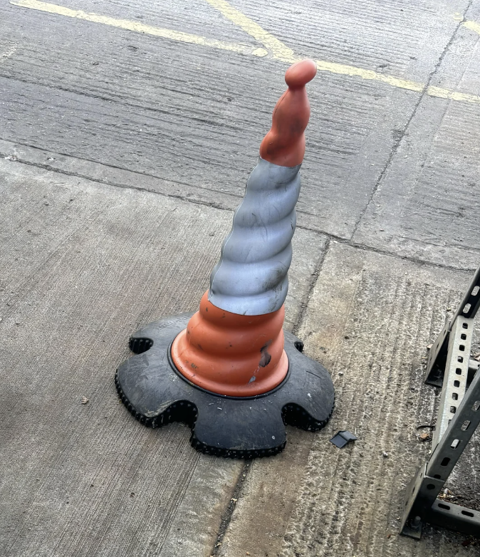 A traffic cone with an irregular, spiraling shape is placed on the ground next to a metal shelf outdoors