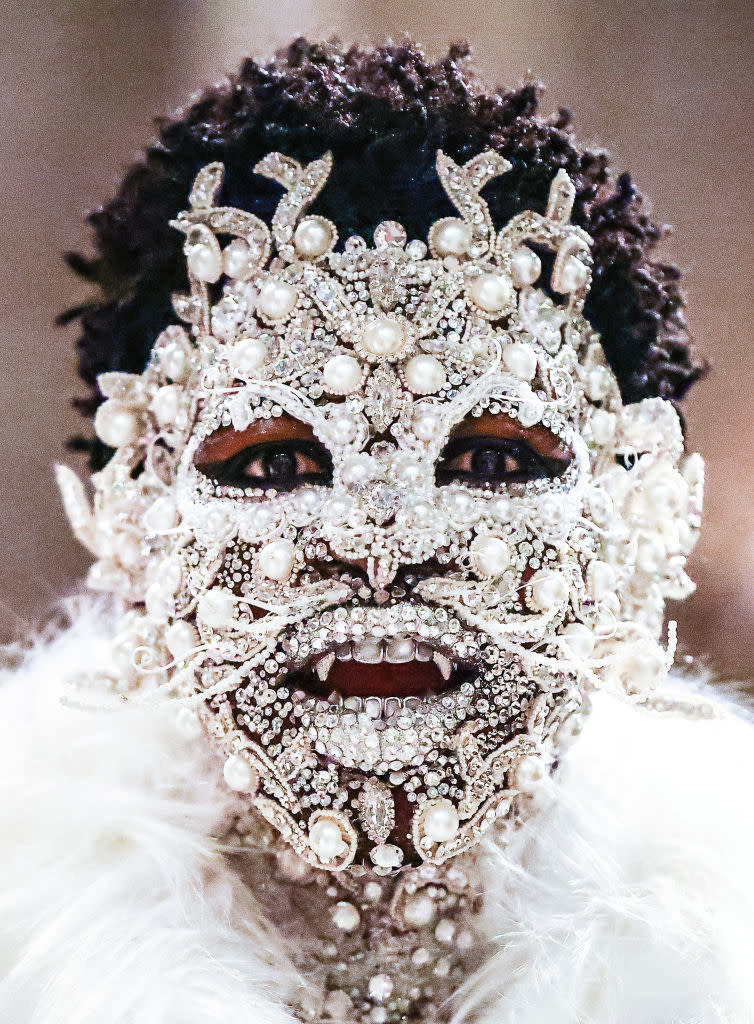 Lil Nas X in a bejeweled mask and fluffy attire. Facial features obscured; expressive eyes visible