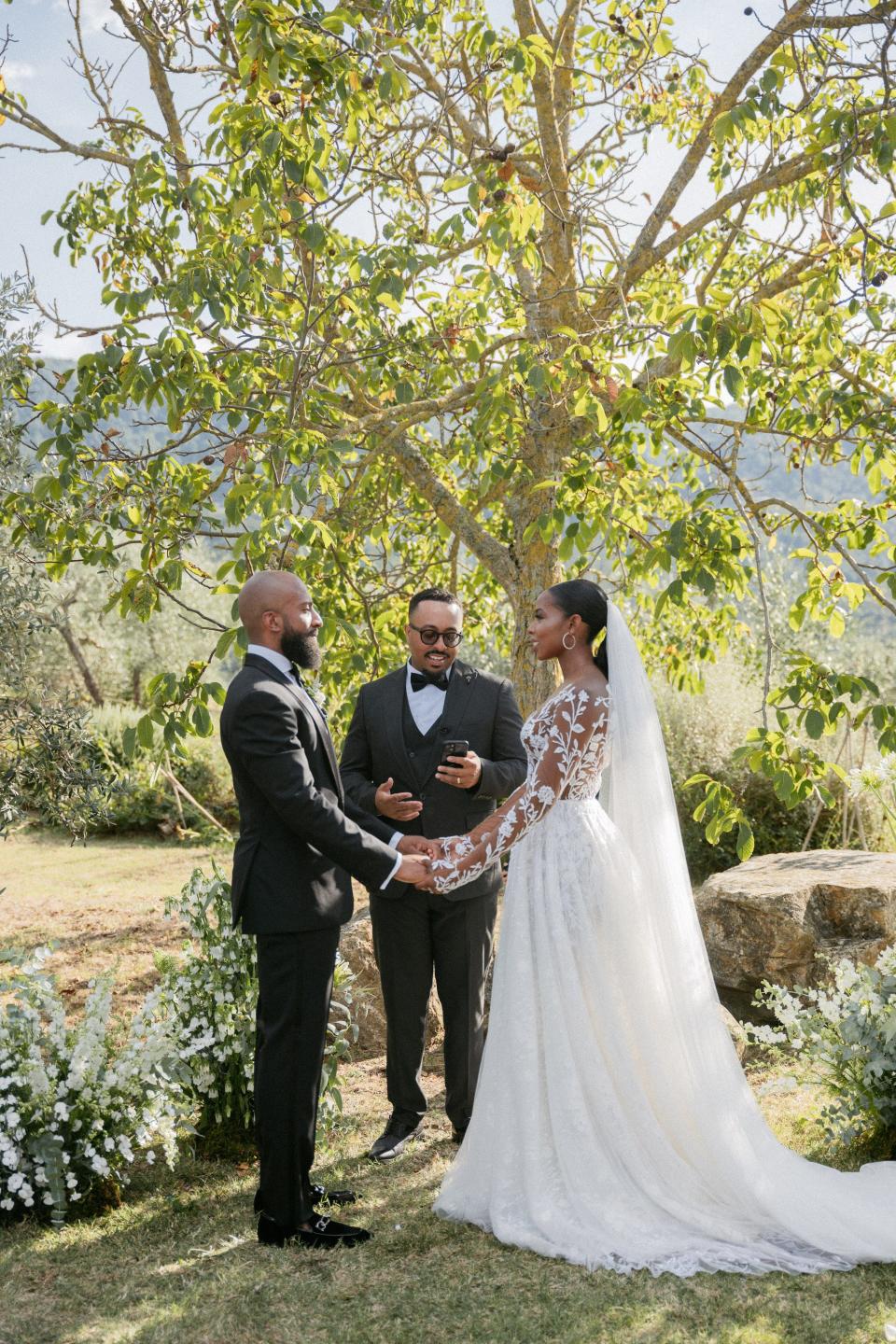 A bride and groom hold hands as they get married in front of trees.