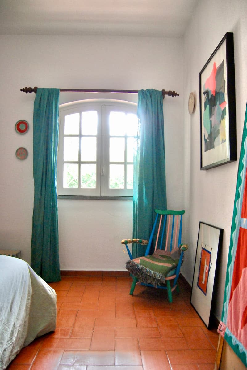 Teal curtains hung on window above colorful chair in white bedroom with rust floor.