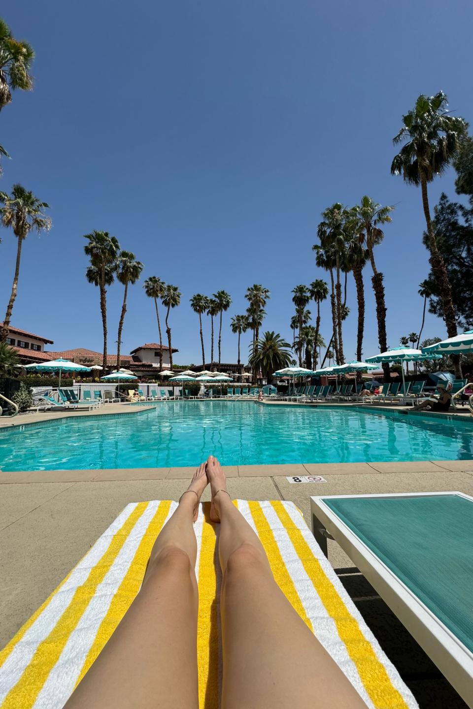 Person relaxing by a pool with outstretched legs, surrounded by palm trees and sun loungers