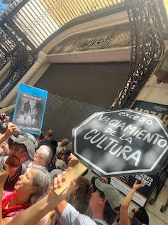 Protestors at holding aloft a black octagonal sign reading 'Exceso Vaciamiento de la Cultura'.