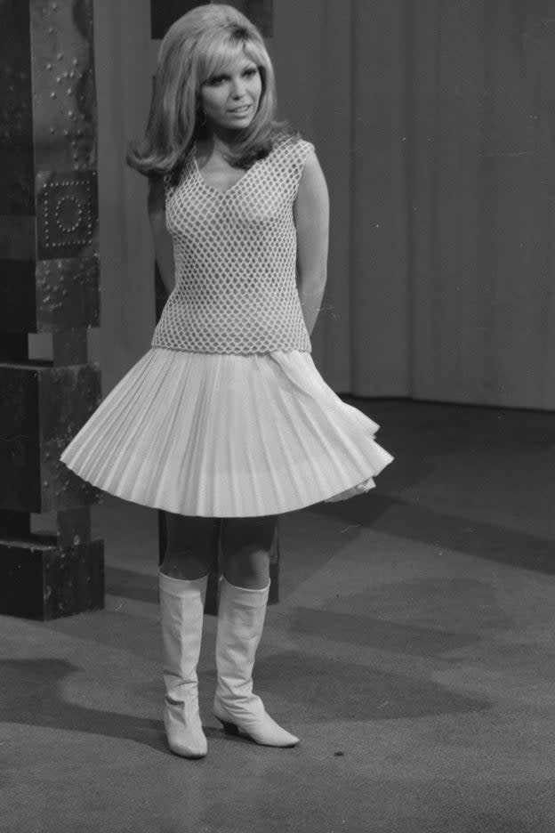 Nancy Sinatra, in a vintage photo, stands with a pleated dress and white boots