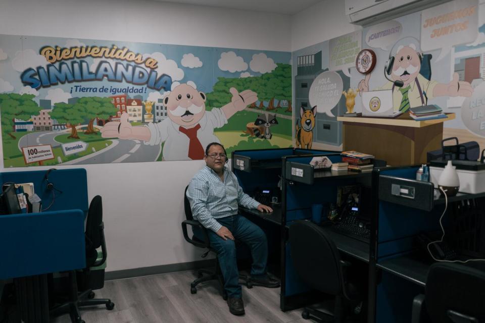 A man sits at a workstation. Behind him is a poster of a mustachioed character and the words Bienvenidos a Similandia