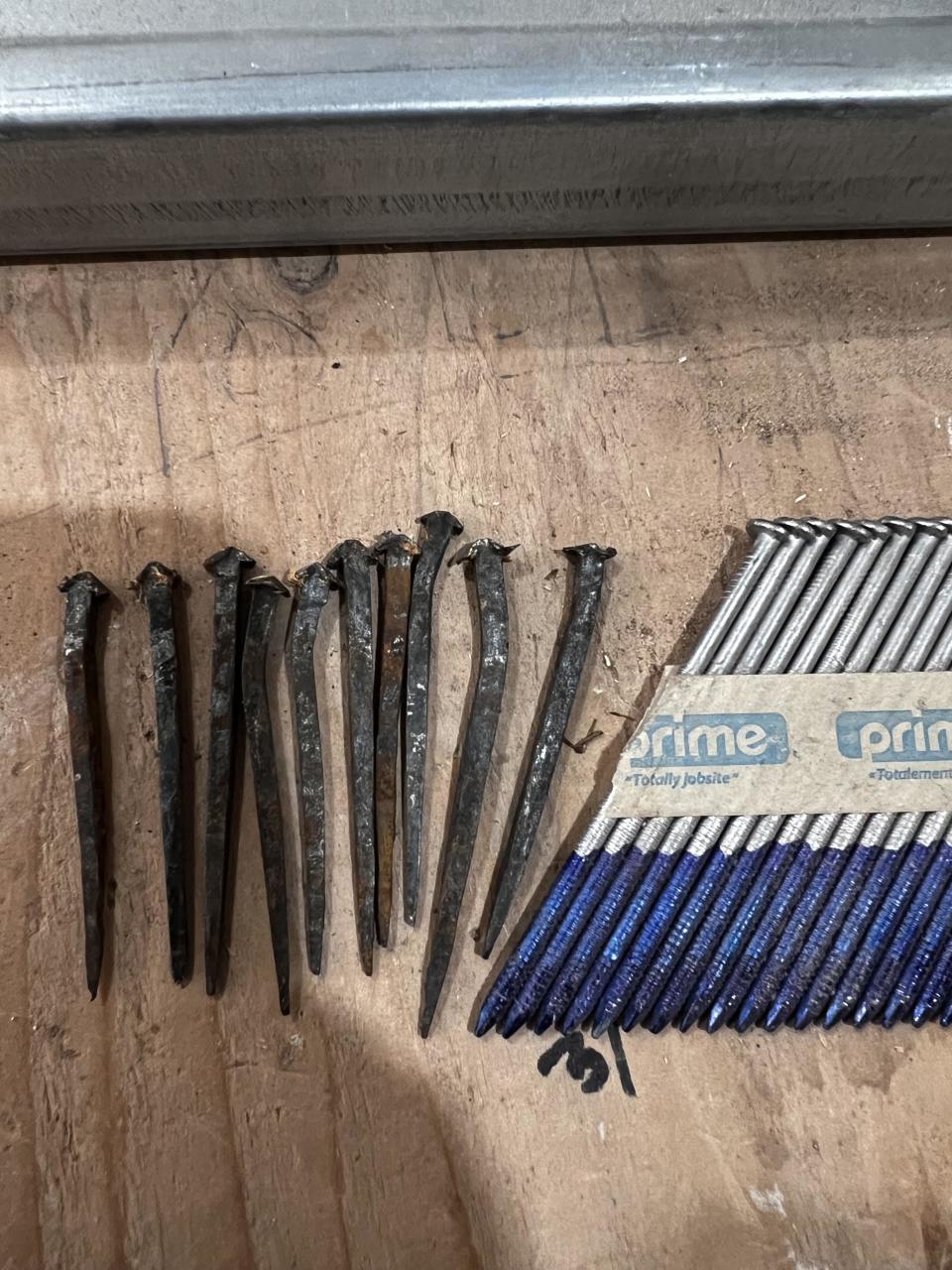 Rusty nails are arranged on a wooden surface next to a bundle of blue and silver nails in Prime packaging