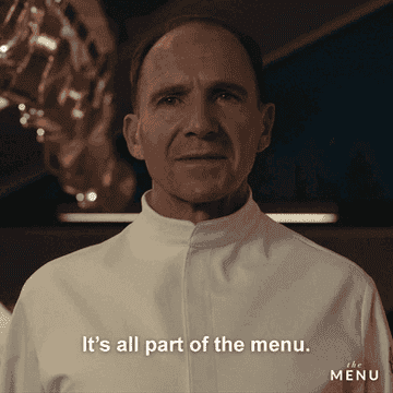 Ralph Fiennes in a white chef's coat, saying, "It's all part of the menu," with "The Menu" logo in the bottom right corner