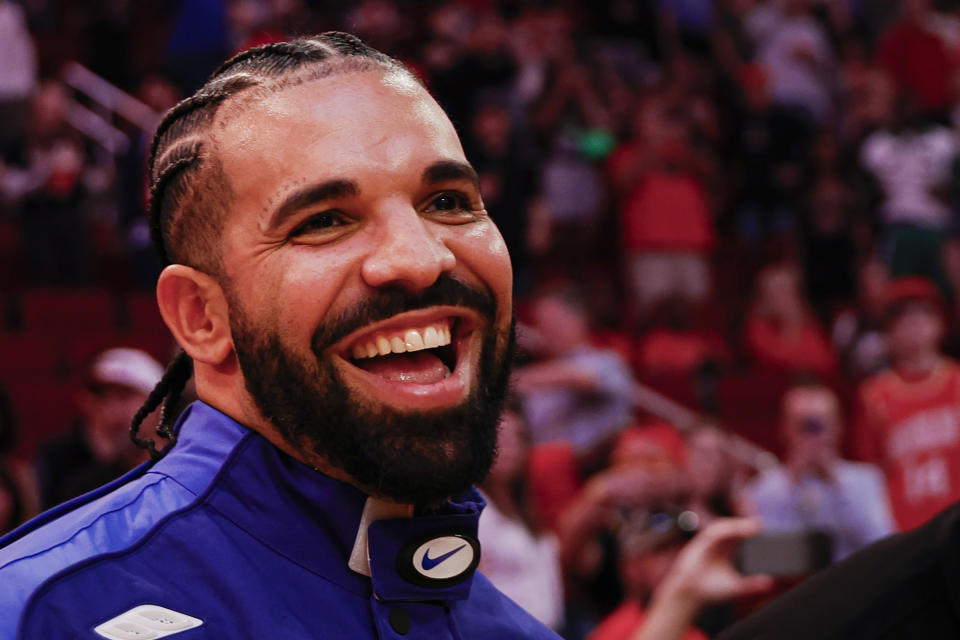 Drake, in a Nike jacket, smiling widely in a crowd at an event