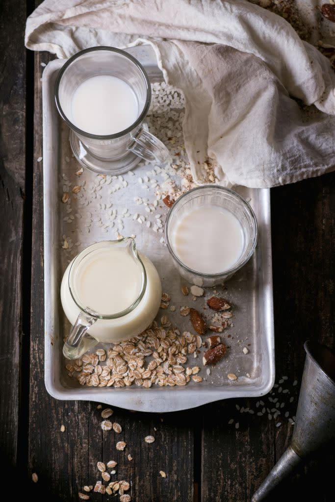 set of non dairy milk rice milk, almond milk and oat milk in glass cups and jug on old aluminum tray with rice grains, oat flakes and almond over old wooden table dark rustic style top view