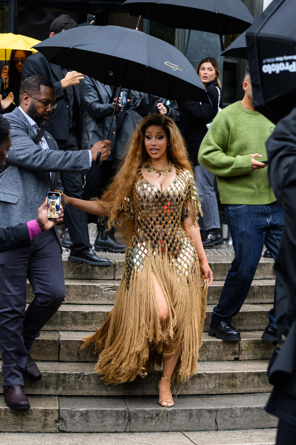 Cardi B in a glamorous gold fringe dress is escorted down steps while holding a black umbrella