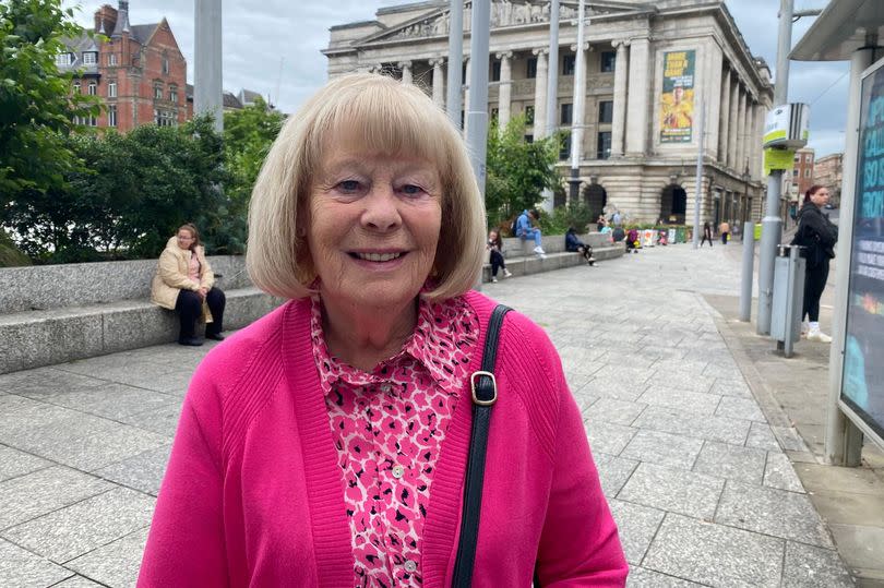 Mrs Watt stood in Old Market Square waiting for her tram smiling at the camera. She has short blonde hair, blue eyes, and is wearing a pink outfit