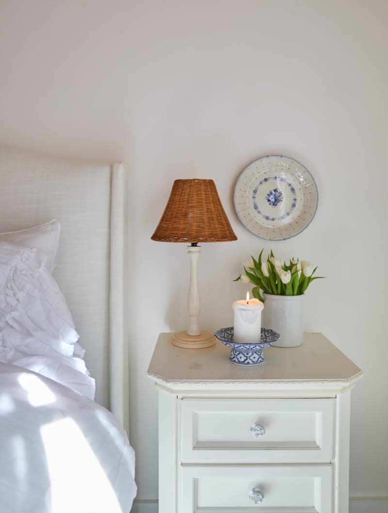 A nightstand with a lamp, candle, and flowers flank a bed with white sheets.