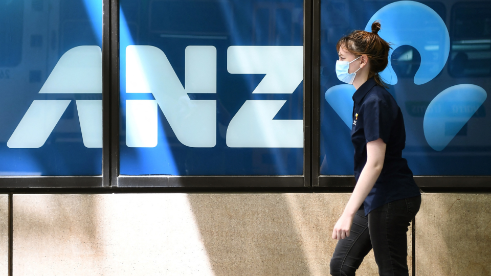 The ANZ logo on the exterior of a building with a woman walking by wearing a mask.