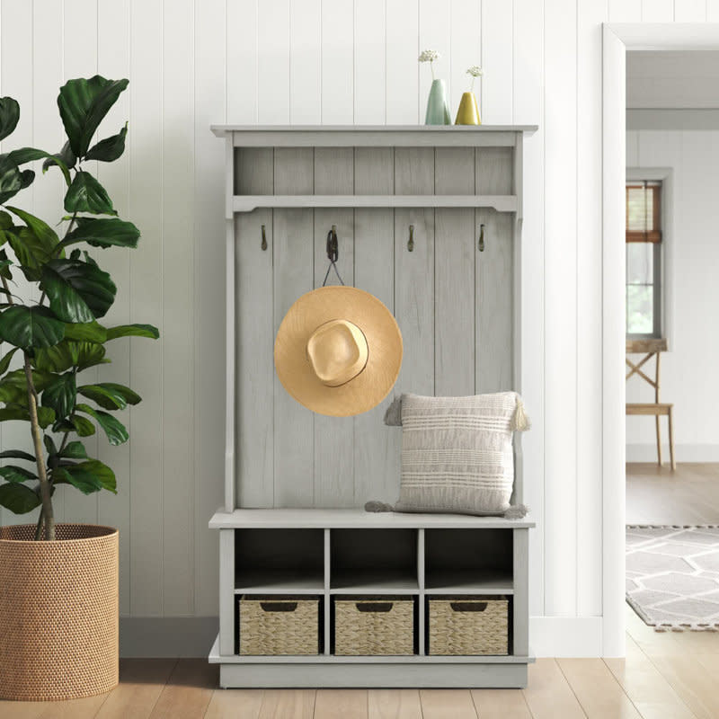 A modern wooden hall tree with hooks for hanging items, a storage bench with cushions, and baskets below, placed next to a plant