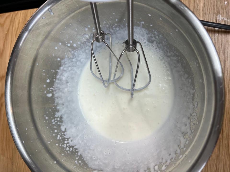 An overhead view of a hand mixer at work in a silver mixing bowl, blending heavy whipping cream and confectioner's sugar.