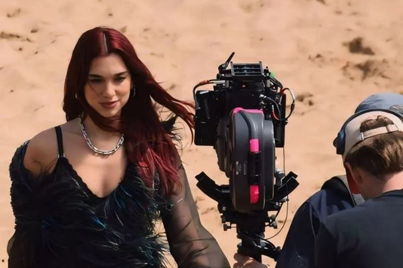 Dua Lipa wearing all black, feathers and red hair with a camera man