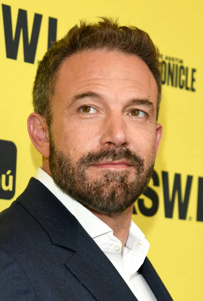 Ben Affleck at an event. He wears a jacket over a white shirt and looks to the left against a yellow background.