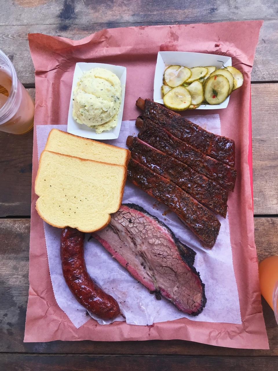 A plate of barbecued meat and side dishes of mashed potatoes, bread, and pickles