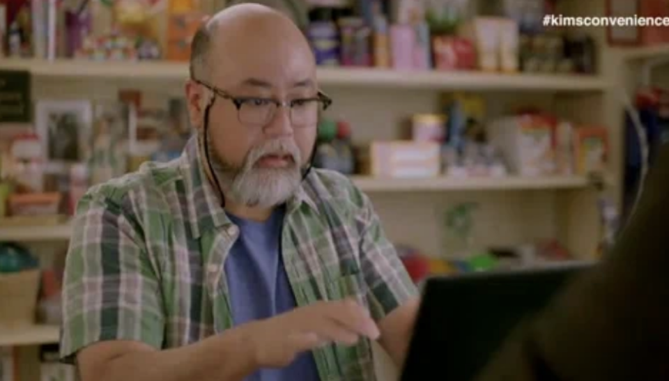 Mr. Kim from Kim's Convenience works at a computer in the store, caption reads 