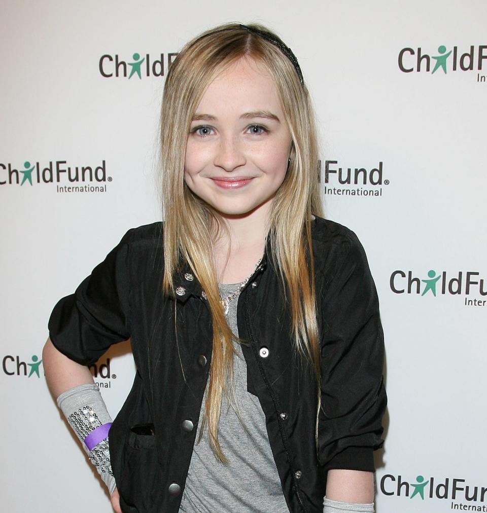 Sabrina Carpenter as a kid in a dark jacket, shirt, frilly black skirt, and patterned tights, standing on the red carpet