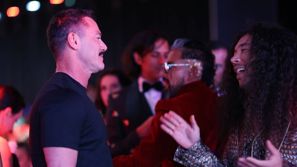 Cole Walliser speaking to actor Luke Evans on a red carpet in Cannes, France, in 2023. Cole (right) is a Canadian man in his 40s with long curly brown hair past his shoulders. He has brown eyes and a short moustache. He has his arms open towards Luke, a white man in his 40s with cropped brown hair and a thick moustache. Cole wears a black and white patterned shirt while Luke is styled in a black T-shirt. Behind them are other red carpet guests and reporters.