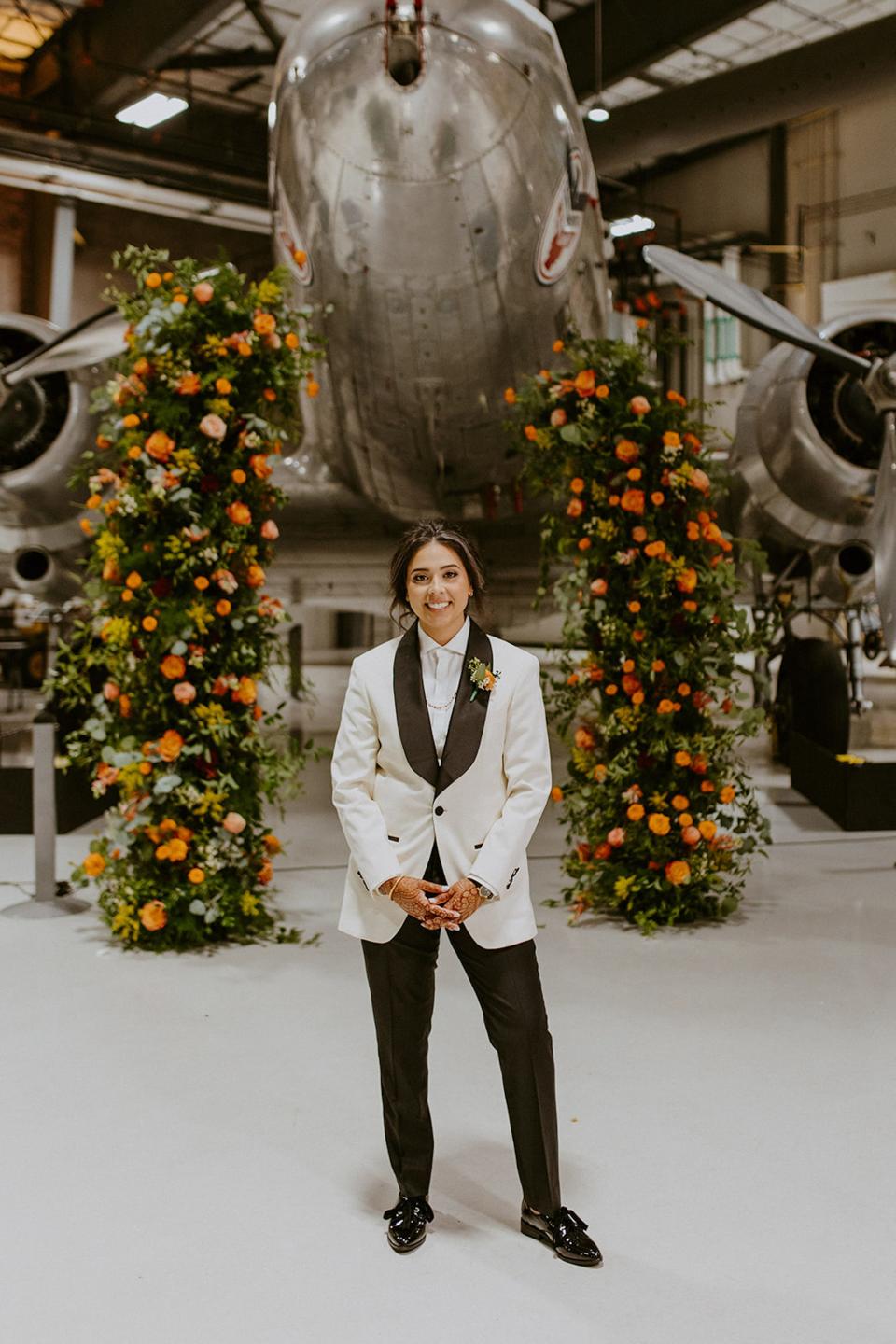 A woman stands in a suit in front of a floral archway and plane.