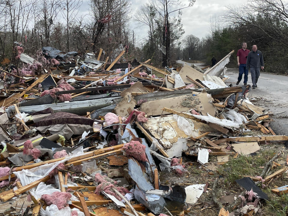 Two people walk along a road next to a pile of debris left after a tornado.