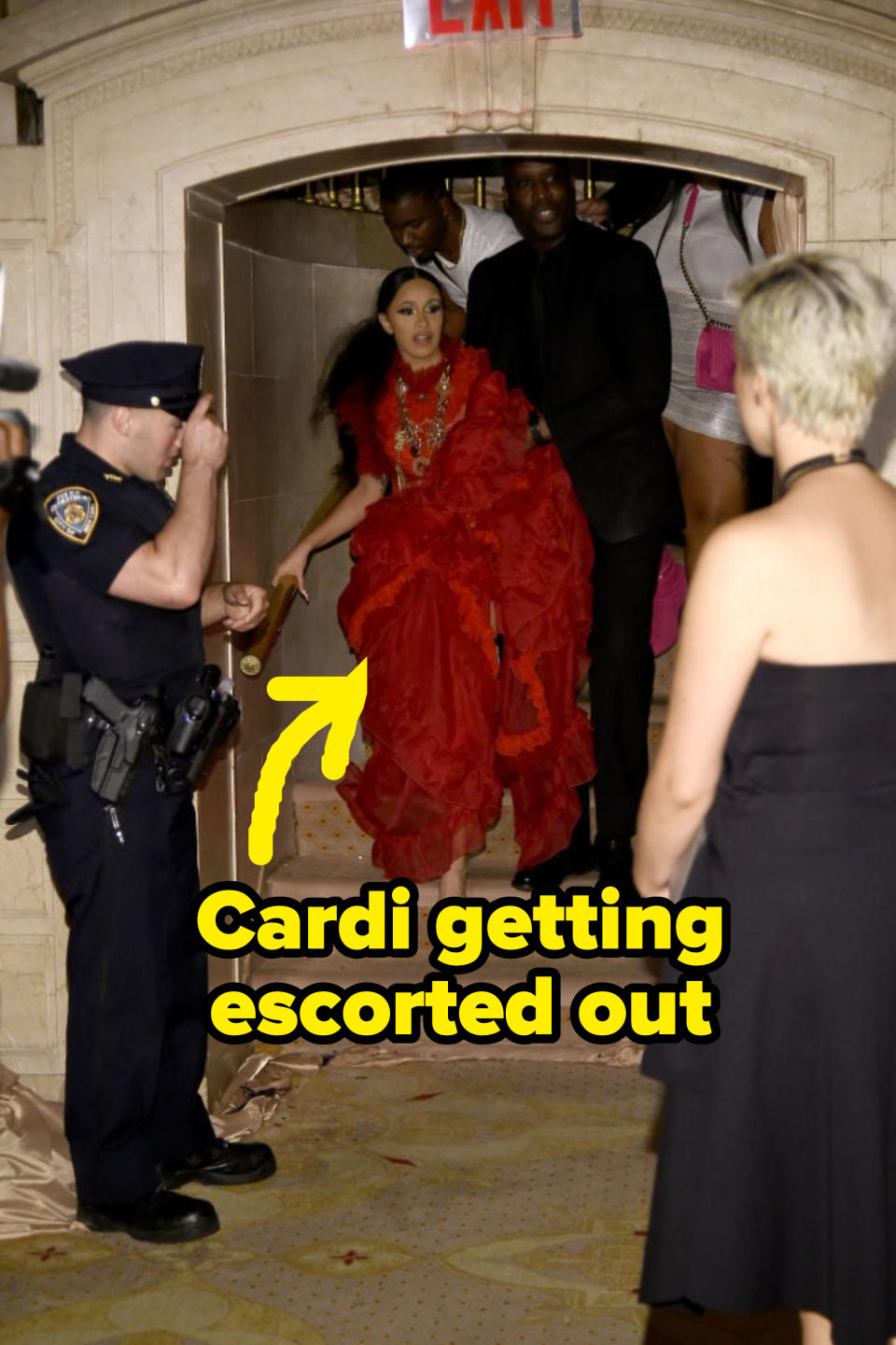 Cardi B in a voluminous ruffled dress with a person, exiting an event as others and security watch