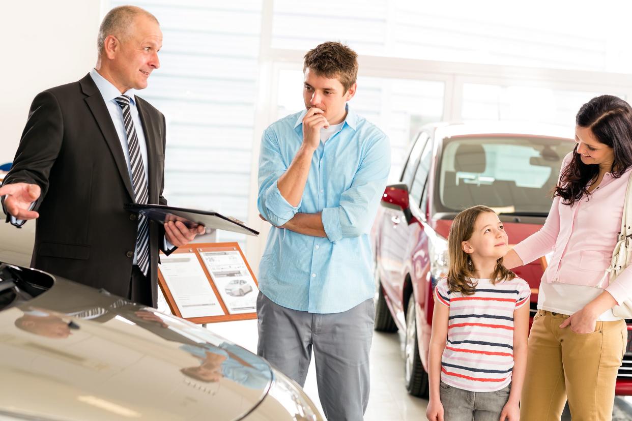 Senior male car dealer offering a vehicle to young family in car dealership showroom, while young man is considering