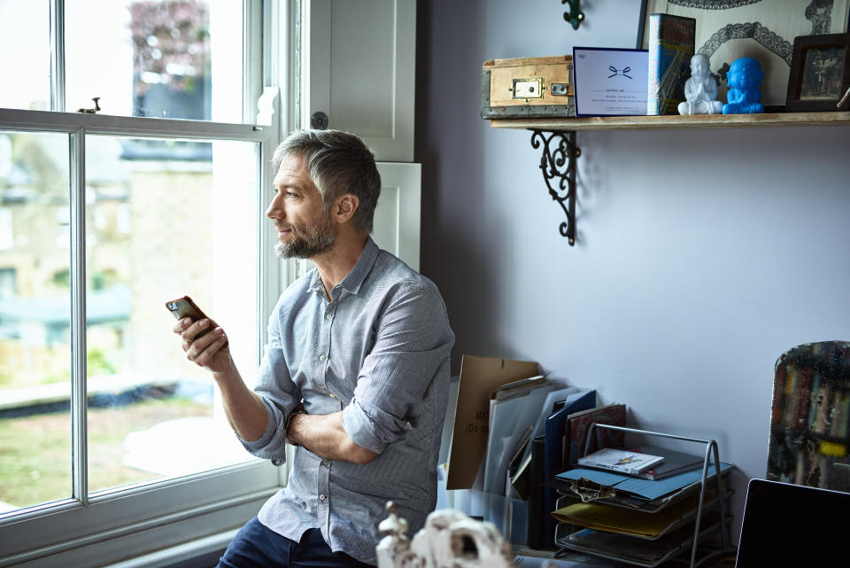 Man in his 40s texting on smartphone, sitting on edge of desk and looking away, contemplation, entrepreneur, small business owner, casual businessman