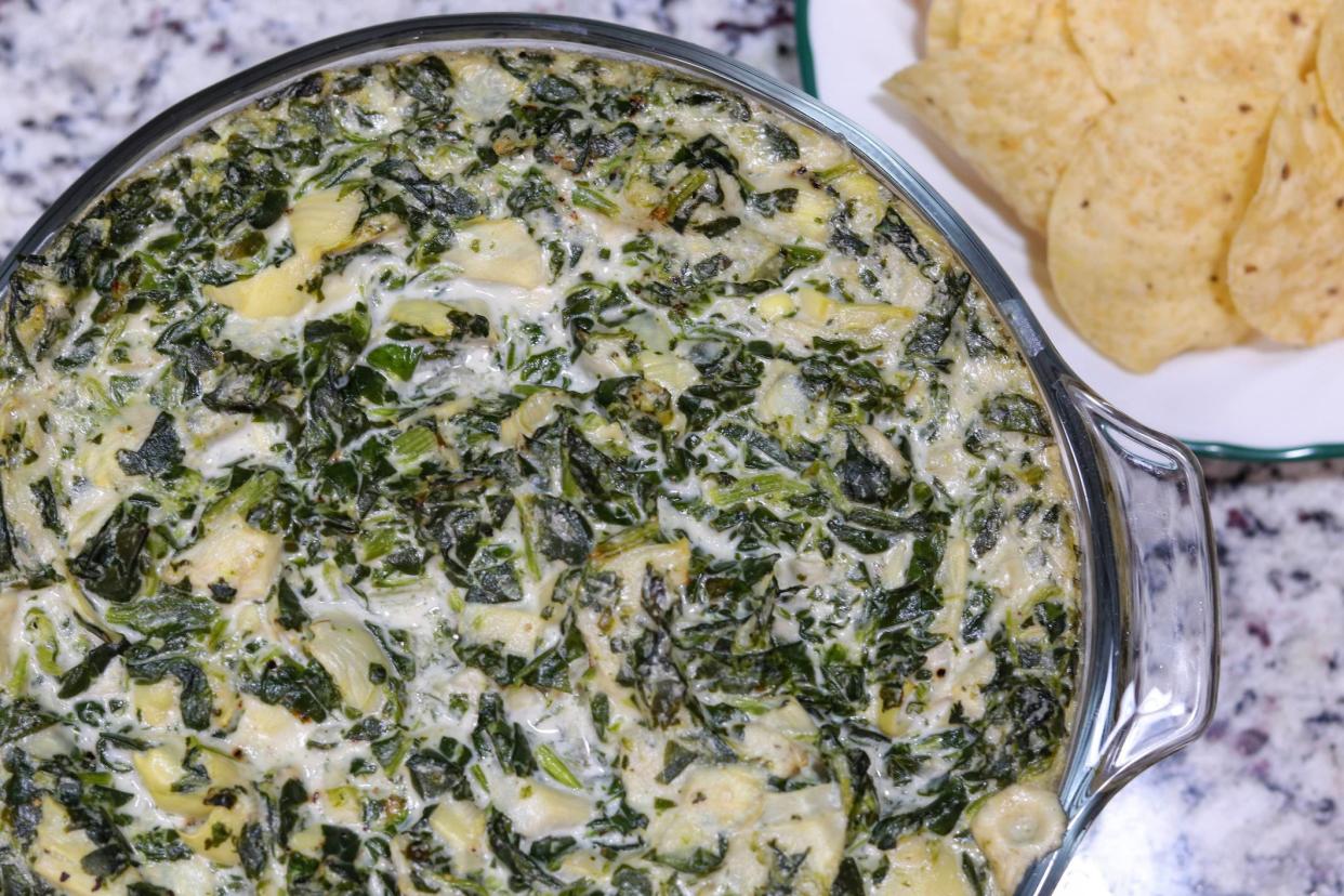 Hot spinach and artichoke dip in a glass bowl with a white plate of tortilla chips on a granite countertop