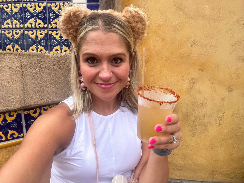 Terri Peters holding a margarita in a glass with a straw. She wears a pair of brown fuzzy ears and a white sleeveless top. Her earrings are shaped like bears. She has shoulder-length blond hair and pink nails. She's posing in front of a yellow wall. Behind her to her right are blue-yellow-and-white tiles.